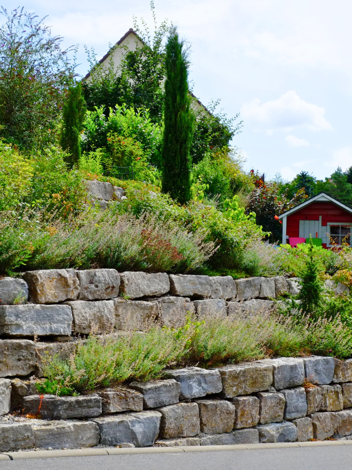 Steinmauer mit grüner Bepflanzung und einem roten Haus im Hintergrund.